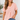 A woman with long, wavy hair is smiling and standing in a bright, minimally decorated room, wearing the Unbranded/Unknown Women’s Pink Short Sleeve V-Neck Blouse - Light Pink with two buttons, paired with blue jeans.