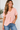 A woman with long, wavy hair is smiling and standing in a bright, minimally decorated room, wearing the Unbranded/Unknown Women’s Pink Short Sleeve V-Neck Blouse - Light Pink with two buttons, paired with blue jeans.