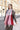A person wears a Women’s Oversized Plaid Winter Blanket Scarf Pink, Black & Red in an oversized, fringed red, pink, and white plaid pattern over a textured corduroy quarter-zip pullover.